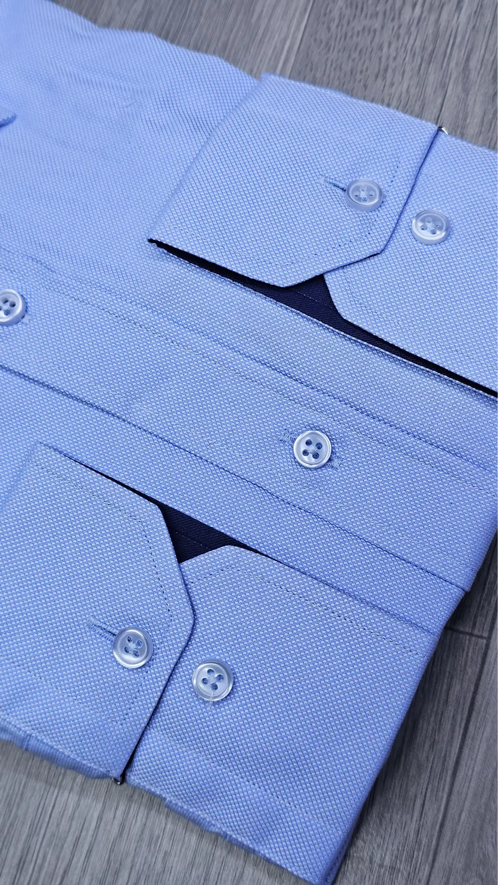 Close-up of a light blue textured dress shirt laid flat on a gray wood-grain surface, showing folded cuffs, buttoned collar, and fabric texture.