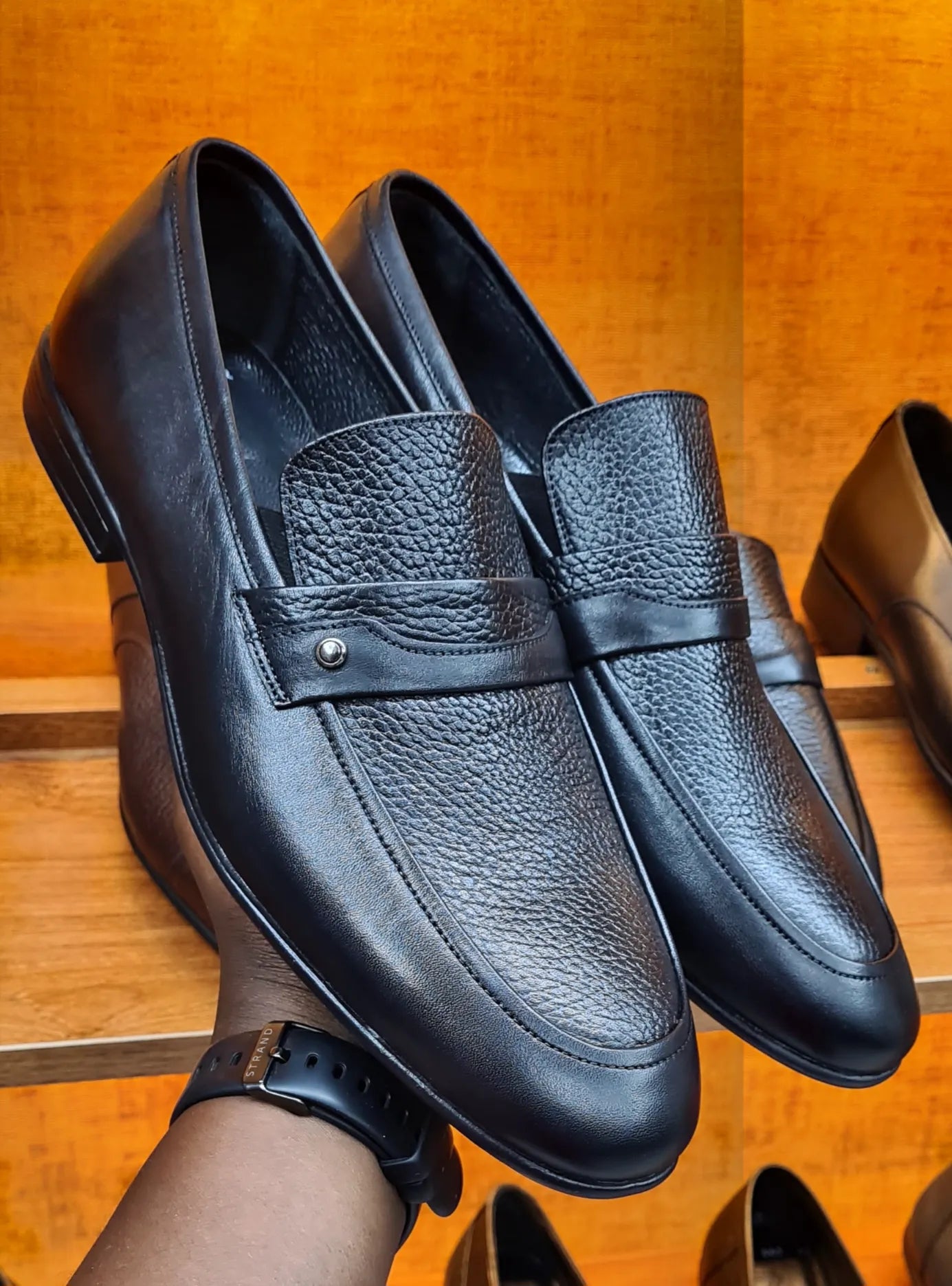 Black leather loafers held by a hand on a wooden surface