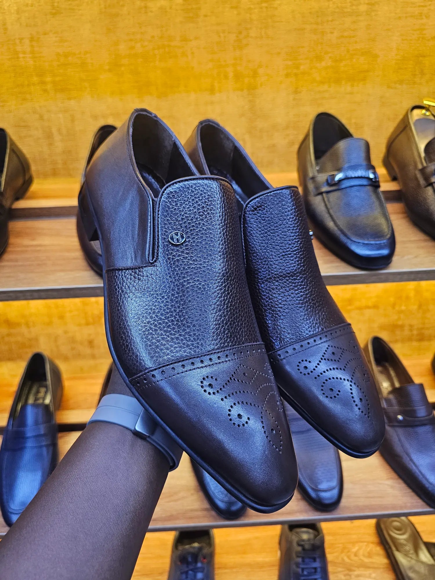 Black leather shoes displayed on a wooden rack with a person holding one shoe.