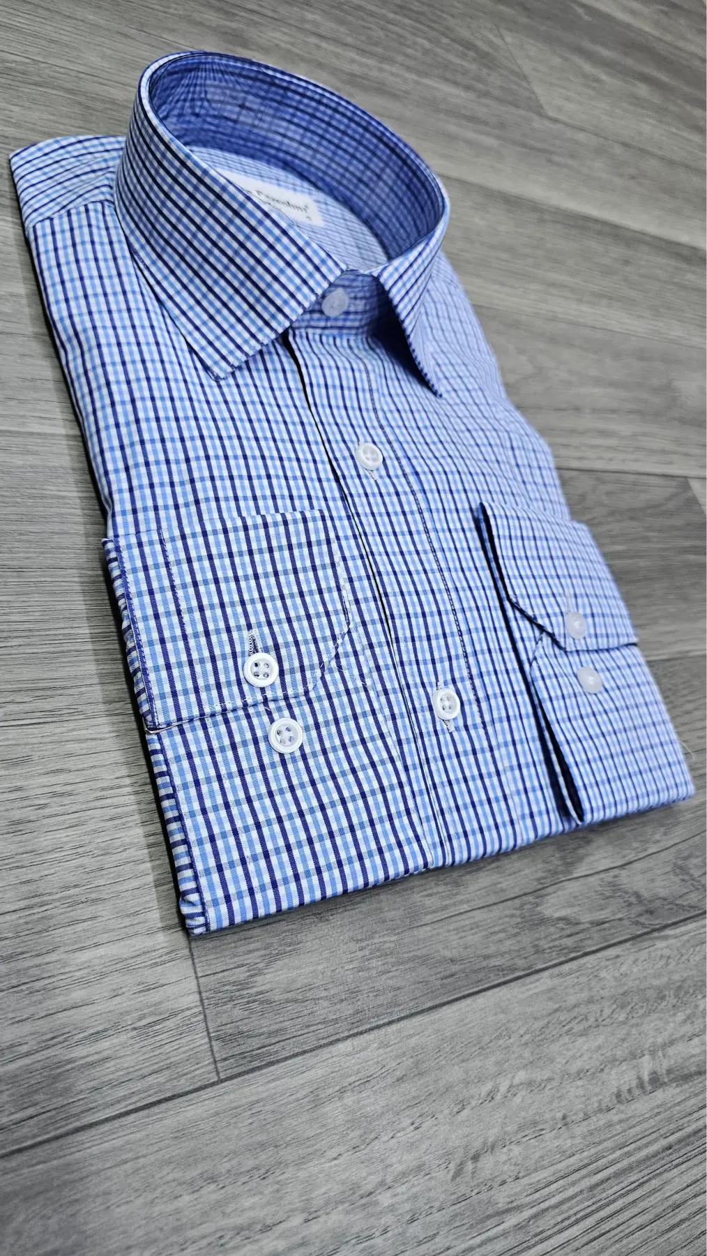 A folded blue and white checkered dress shirt with a pointed collar and white buttons, laid flat on a gray wood-look floor.