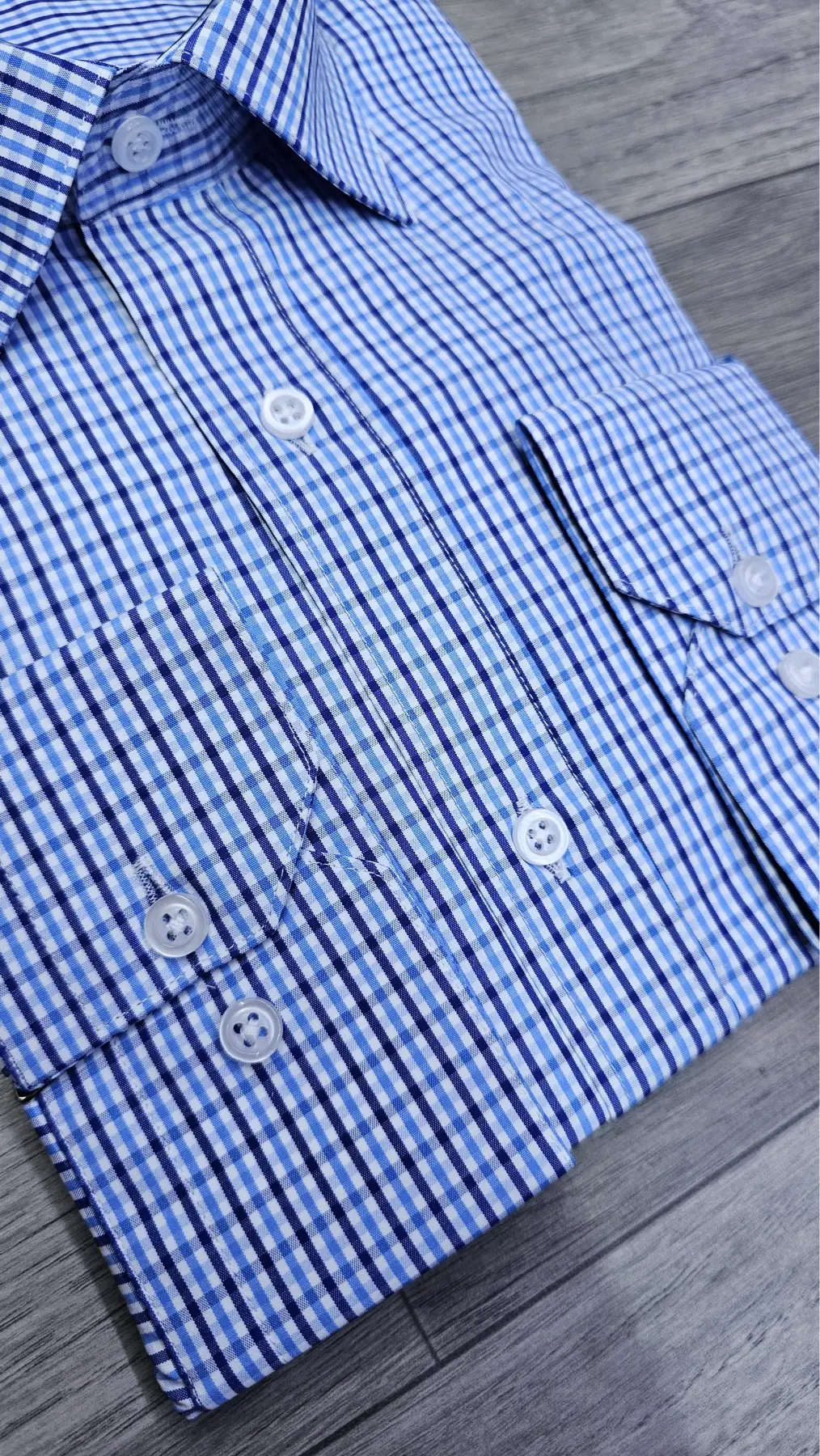 A close-up of a folded blue and white gingham dress shirt with white buttons, laid flat on a gray wood-textured surface.