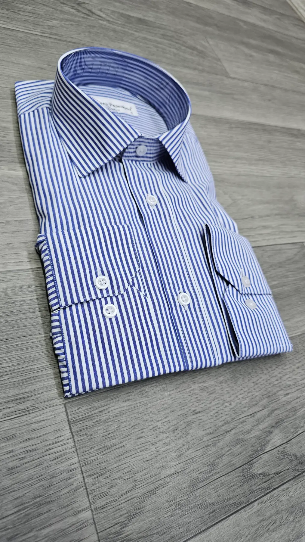 A folded blue and white striped dress shirt with a pointed collar and button-down front, laid flat on a gray wood-look floor.