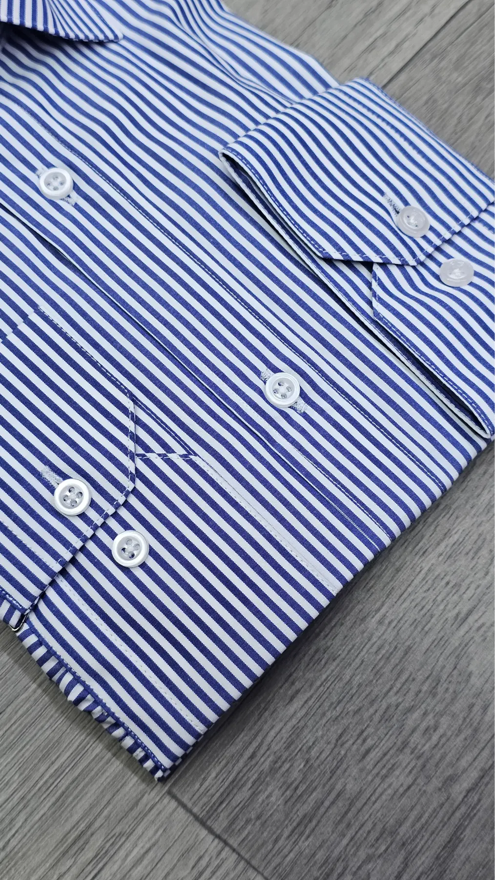 Close-up of a blue and white striped dress shirt folded on a gray wood-textured surface, showing the striped fabric, white buttons, and part of the pocket.