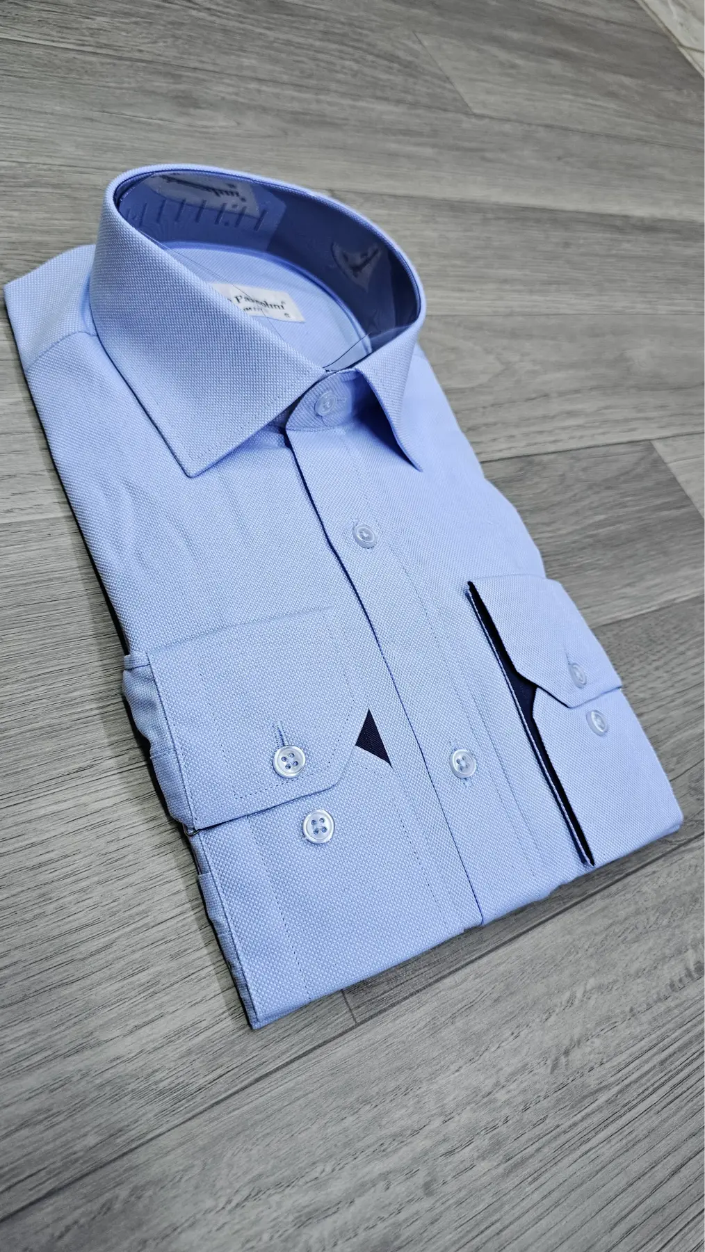 A light blue dress shirt folded neatly on a gray wood-look floor, featuring a spread collar, long sleeves, and a distinctive black triangular accent on the front pocket.