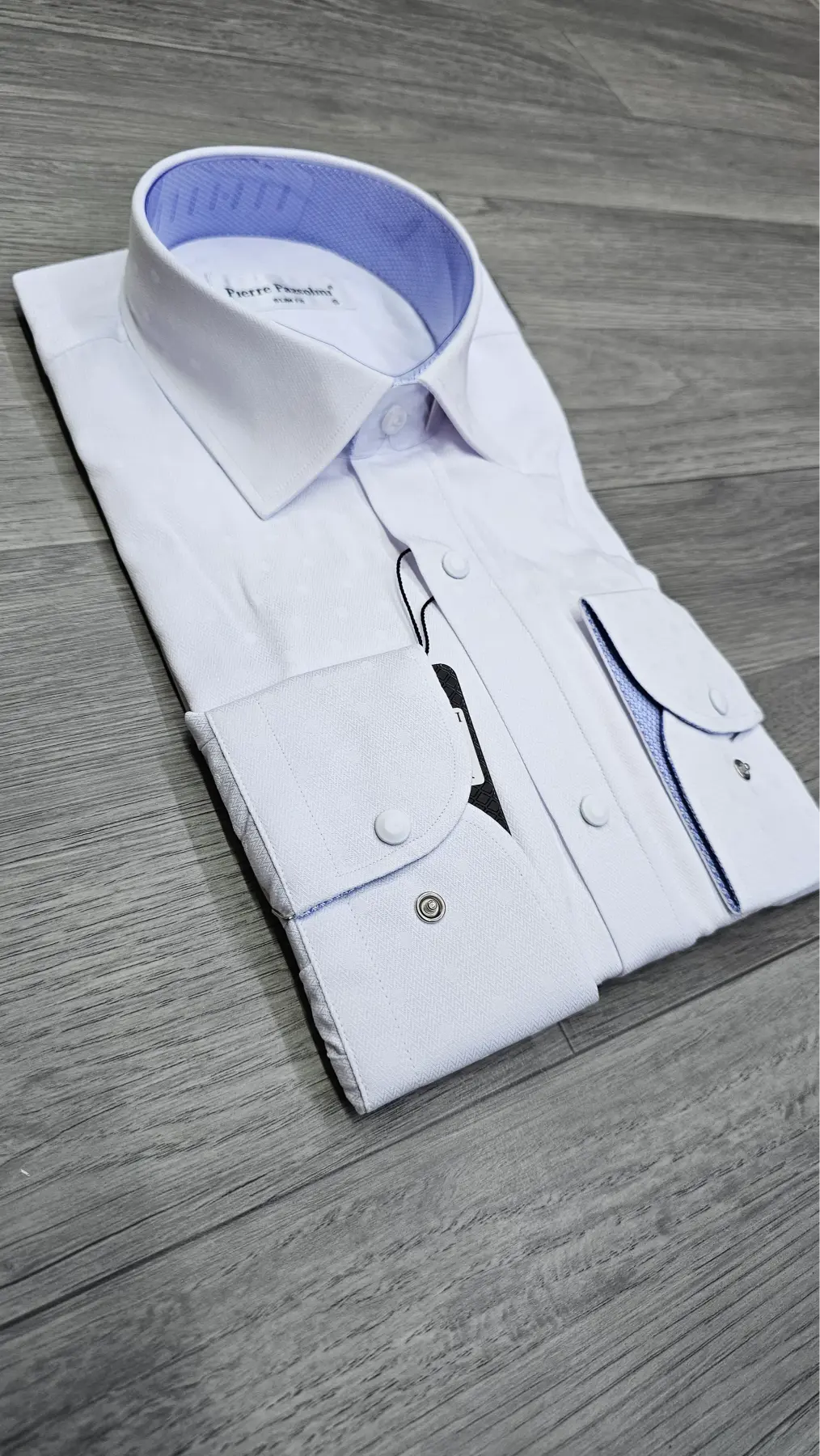 A folded white dress shirt with a light blue inner collar and blue piping details, displayed on a gray wood-textured surface.