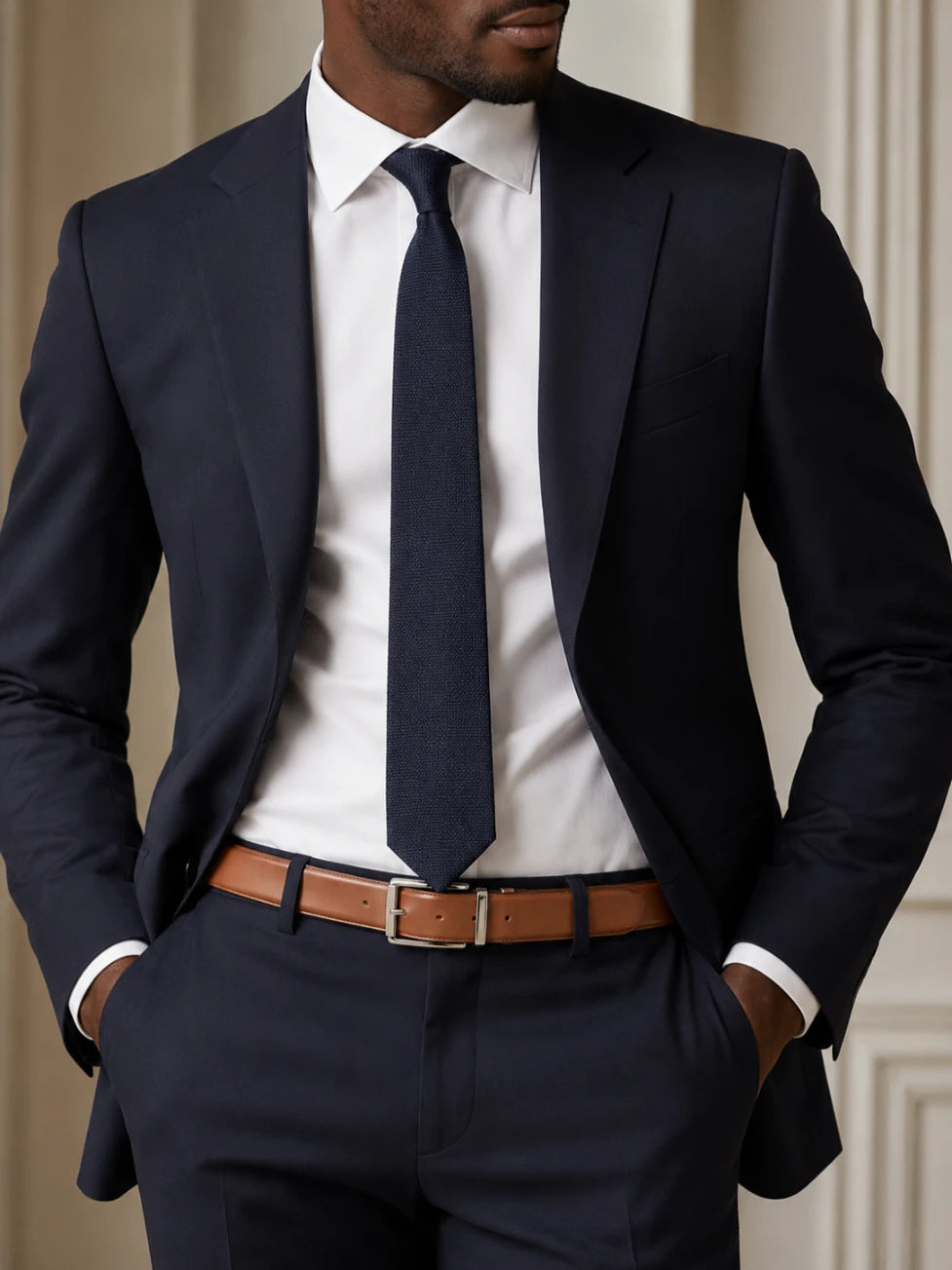 A man wearing a navy blue suit with a white dress shirt, a matching navy tie, and a brown leather belt, shown from the chest down in a clean indoor setting.