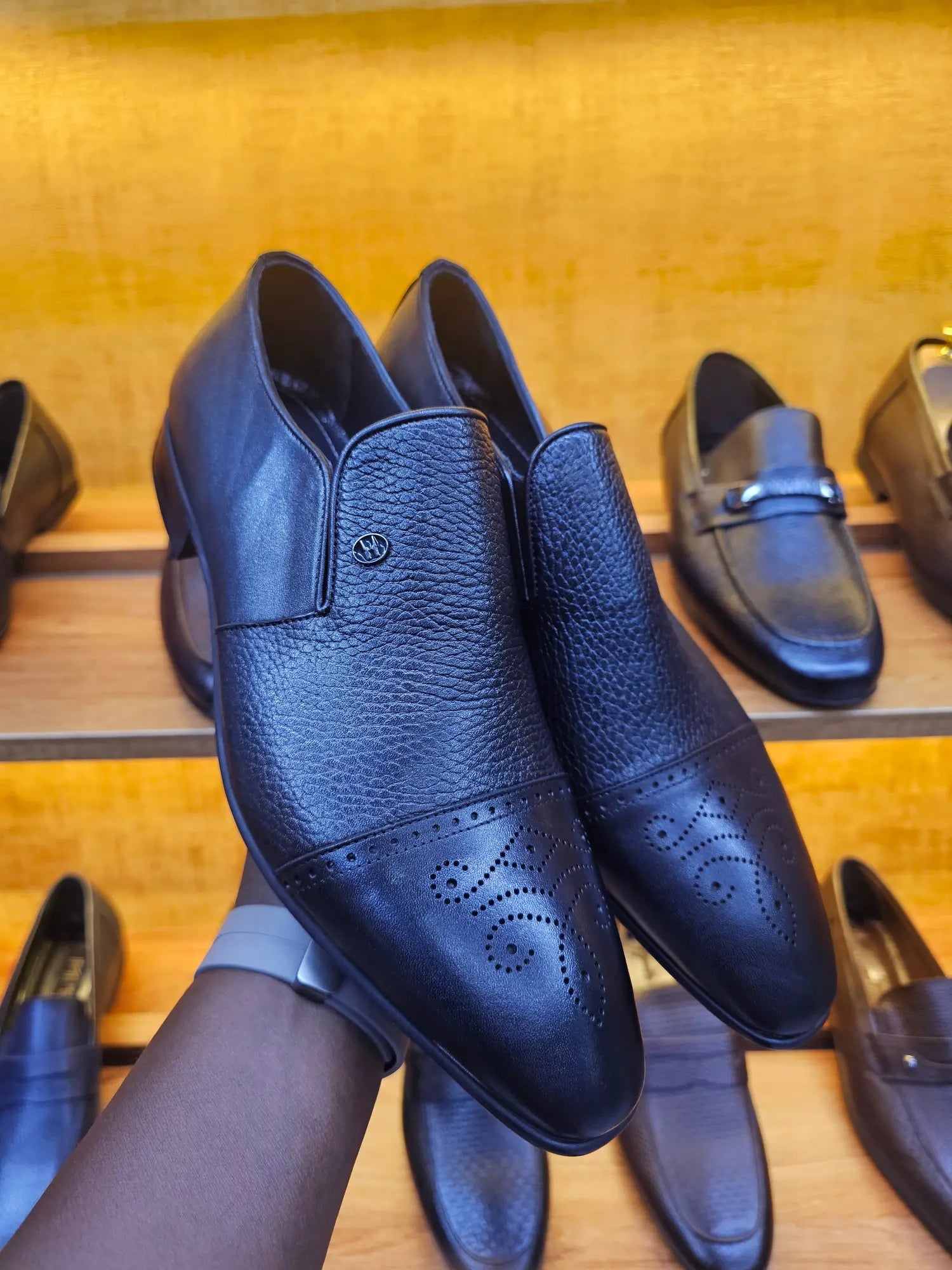 Black leather shoes being held against a wooden background with other shoes displayed.