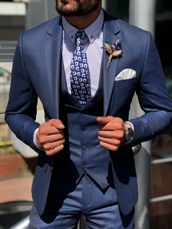 Man wearing a blue suit with a patterned tie and boutonniere, standing outdoors.