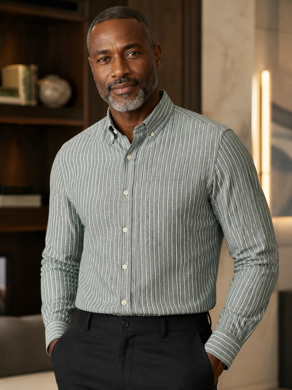 A man wearing a jungle green and white vertically striped long-sleeve button-down shirt, paired with black trousers, standing in a warmly lit indoor setting.