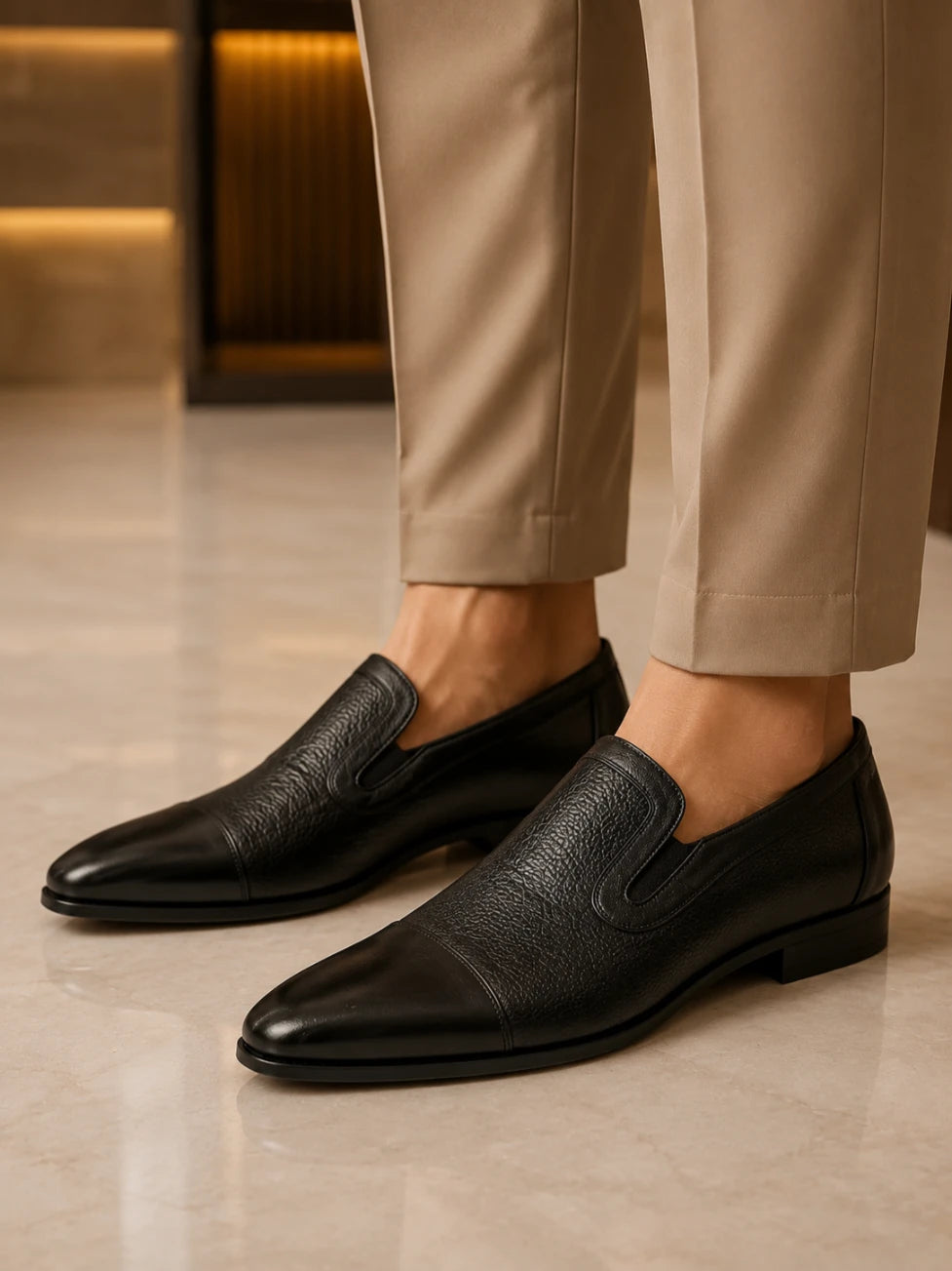 Black leather loafers worn with beige pants on a light wooden floor.