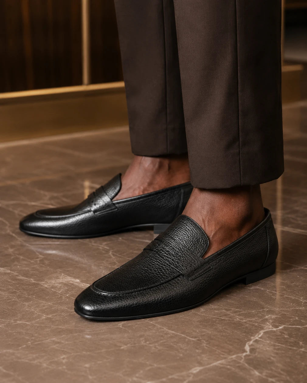 Black leather loafers worn with brown pants on a marble floor.