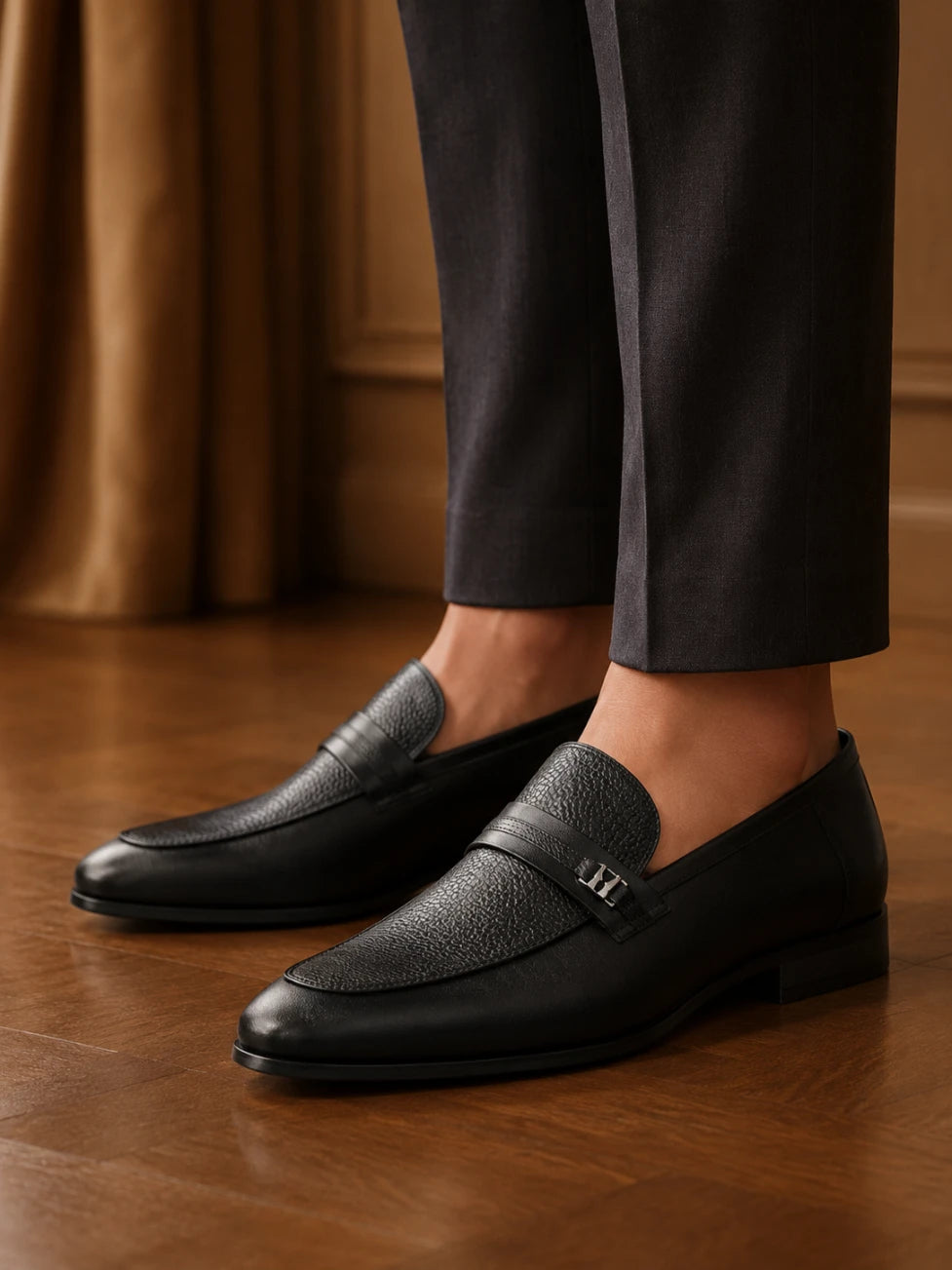 Black leather loafers worn with dark trousers on a wooden floor.