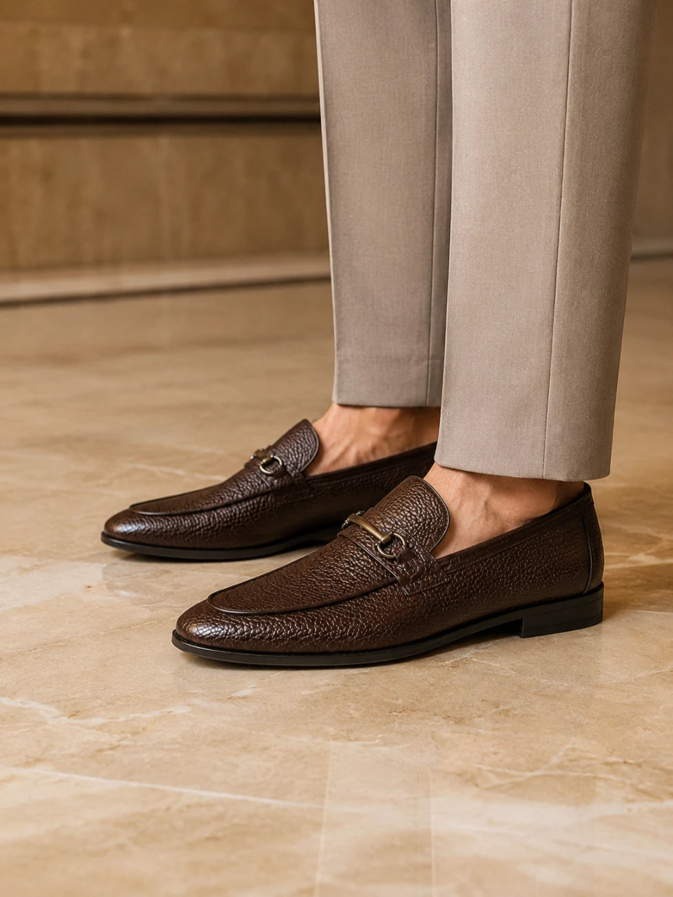 Brown leather loafers worn with beige pants on a marble floor.