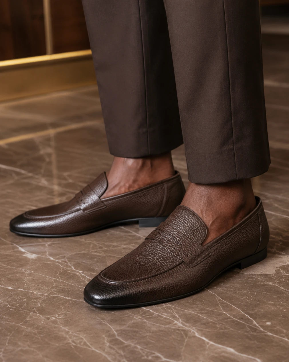 Brown leather loafers worn with brown pants on a marble floor.