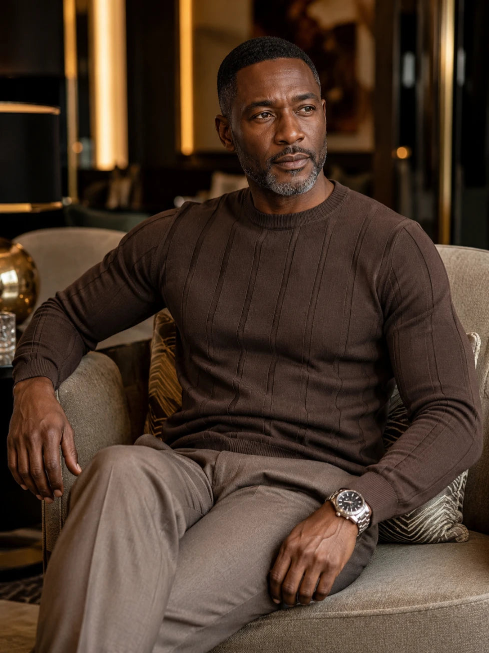 Man sitting on a couch wearing a brown sweater and beige pants in a dimly lit room.