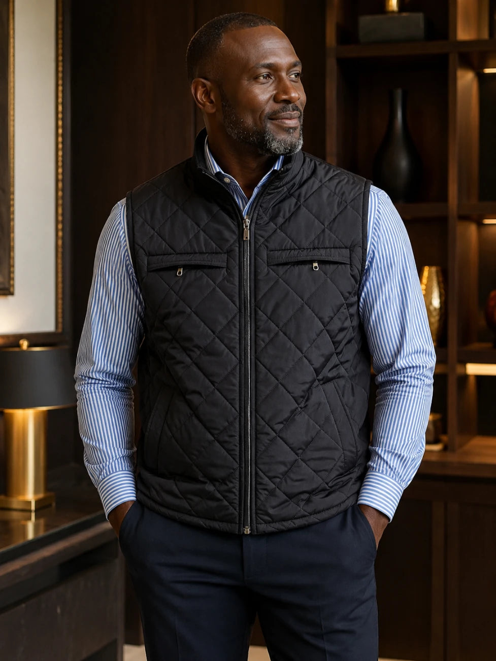 Man wearing a black quilted vest over a blue striped shirt in an indoor setting.