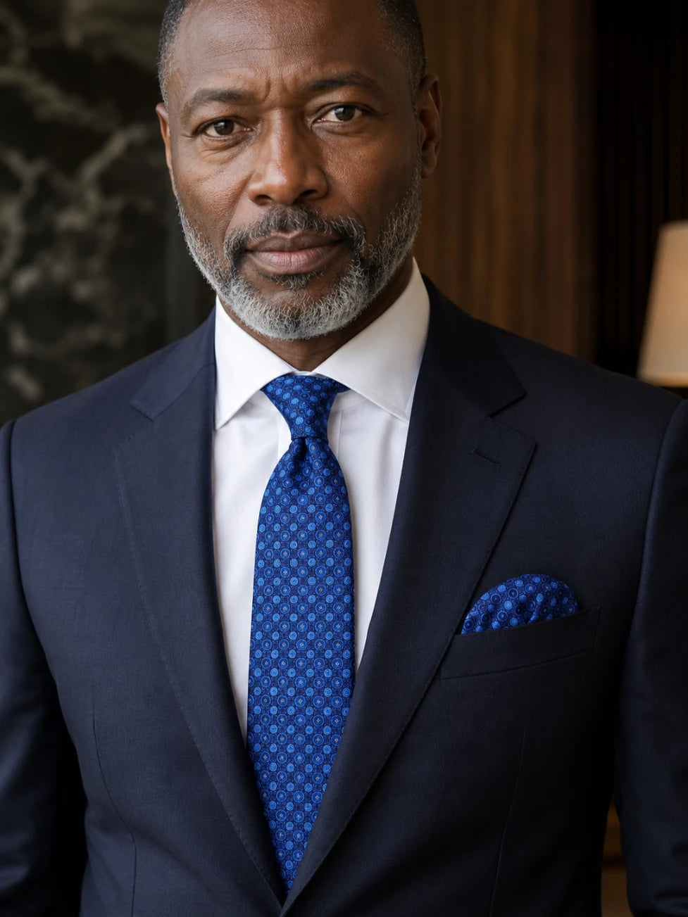 Man wearing a dark suit with a blue tie and pocket square indoors.