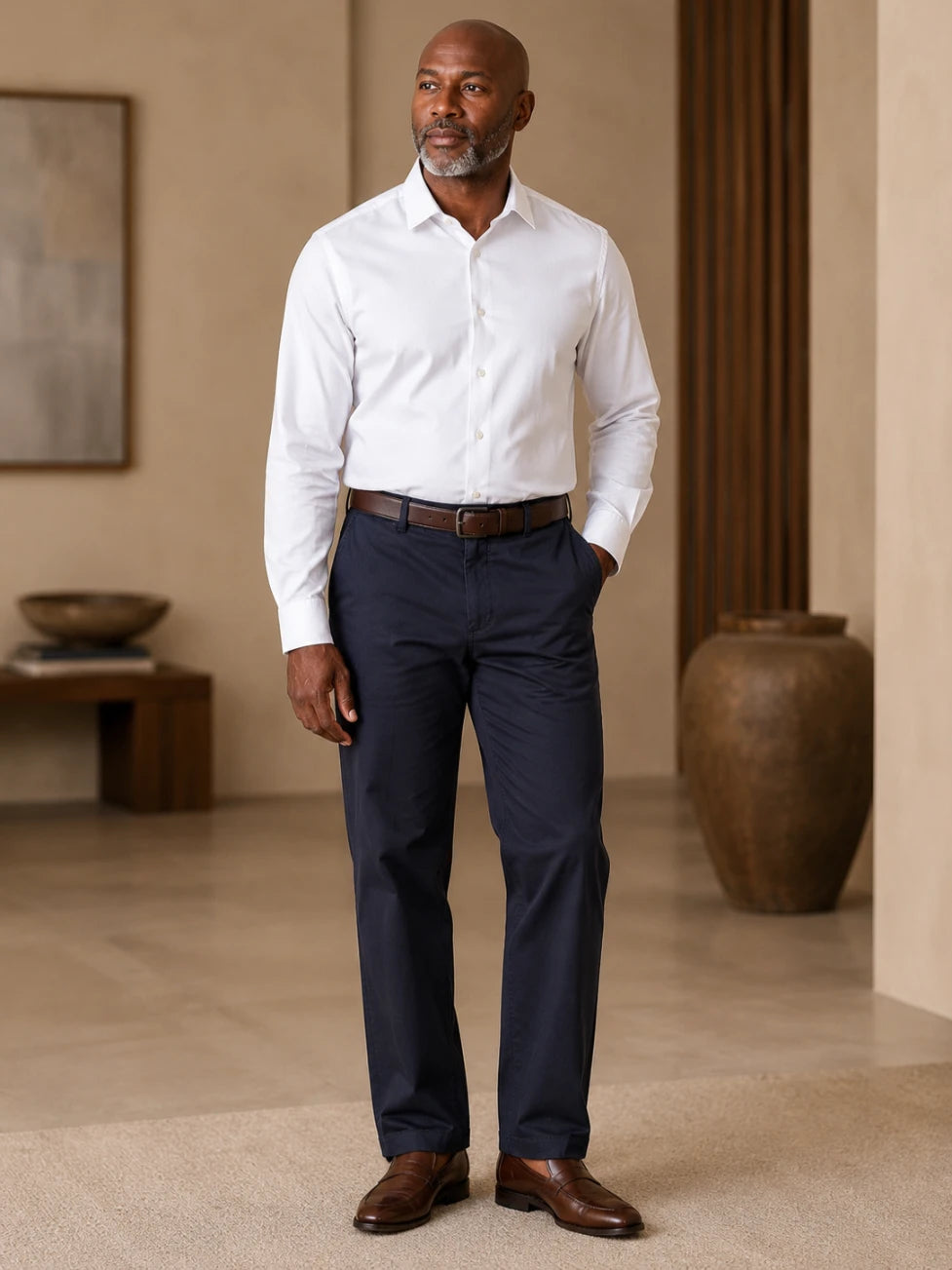 Man wearing a white dress shirt and black pants standing in a room with beige walls and a large vase.